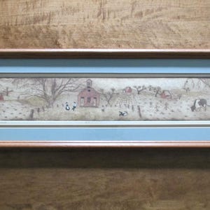 Op de afbeelding: Ingelijste landschapskunstprint met een landelijke scène met een rode kerk, bomen en figuren. Het kunstwerk is geplaatst in een lichtblauwe passe-partout en een bruine houten lijst. De algehele esthetiek is rustiek en charmant.