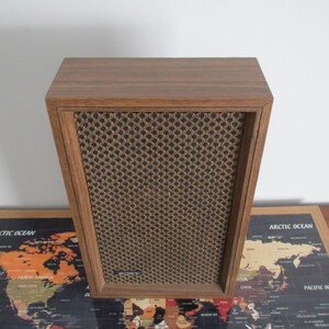 May include: A vintage wooden speaker with a brown finish and a black and tan mesh front. The speaker has a rectangular shape with a slightly angled top. The speaker is sitting on a table with a world map.
