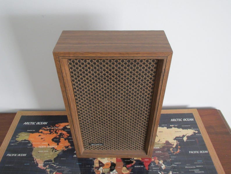 May include: A vintage wooden speaker with a brown finish and a black and tan mesh front. The speaker has a rectangular shape with a slightly angled top. The speaker is sitting on a table with a world map.