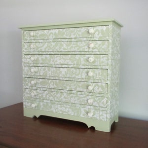 May include: A light green wooden dresser with six drawers and white knobs. The drawers are decorated with a white lace pattern.
