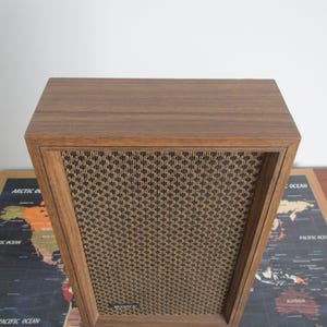 May include: A vintage wooden speaker with a brown wood grain finish. The speaker has a rectangular shape with a woven mesh front panel. The speaker is sitting on a world map.