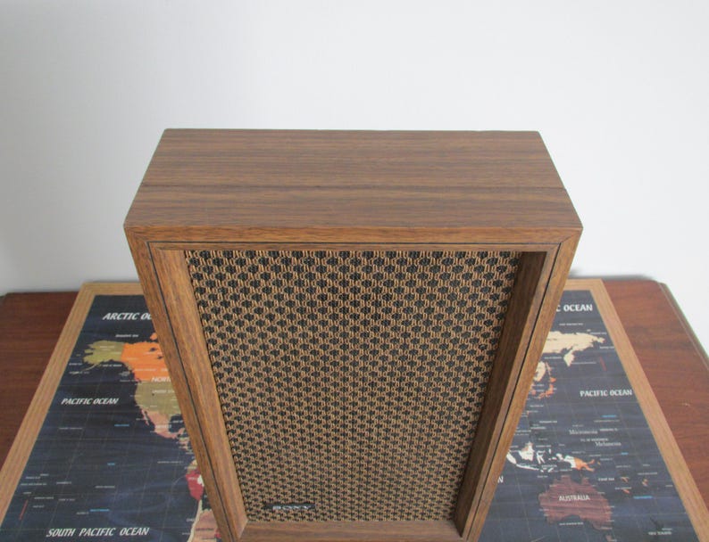 May include: A vintage wooden speaker with a brown wood grain finish. The speaker has a rectangular shape with a woven mesh front panel. The speaker is sitting on a world map.