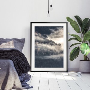 May include: A black framed photograph of a misty forest scene with a light shining through the trees. The photograph is hanging on a white wall in a bedroom setting with a bed and a plant in the corner.