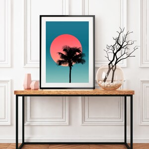Pode incluir: Uma impress&atilde;o emoldurada de uma silhueta de palmeira contra um p&ocirc;r do sol azul e rosa. A impress&atilde;o est&aacute; sobre uma mesa de madeira com um vaso de galhos e dois objetos decorativos cor-de-rosa.