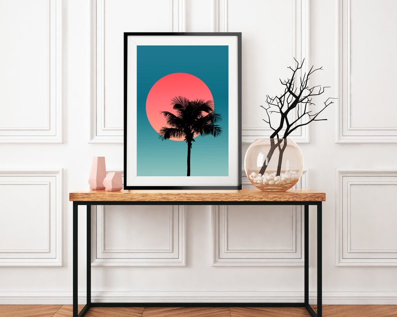 Pode incluir: Uma impress&atilde;o emoldurada de uma silhueta de palmeira contra um p&ocirc;r do sol azul e rosa. A impress&atilde;o est&aacute; sobre uma mesa de madeira com um vaso de galhos e dois objetos decorativos cor-de-rosa.