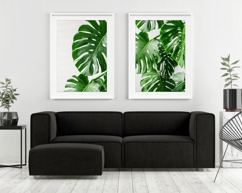 May include: Two framed prints of large green monstera leaves against a white background, hanging above a black sofa and ottoman in a modern living room.
