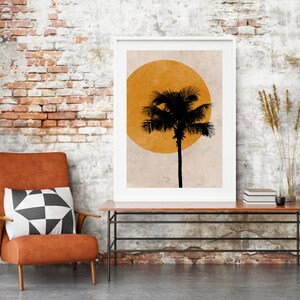 K&ouml;nnte beinhalten: Ein gerahmter Kunstdruck mit einer schwarzen Palmensilhouette vor einer gelben Sonne. Der Druck steht auf einem Holztisch mit Metallrahmen, neben einem Ledersessel mit einem schwarz-wei&szlig;en geometrischen Kissen. Der Hintergrund ist eine Backsteinmauer.