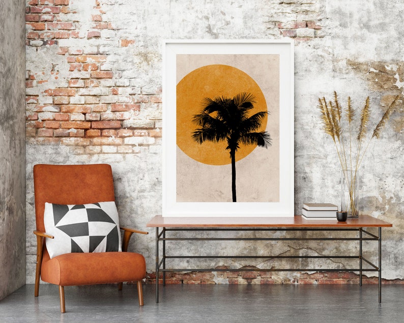 K&ouml;nnte beinhalten: Ein gerahmter Kunstdruck mit einer schwarzen Palmensilhouette vor einer gelben Sonne. Der Druck steht auf einem Holztisch mit Metallrahmen, neben einem Ledersessel mit einem schwarz-wei&szlig;en geometrischen Kissen. Der Hintergrund ist eine Backsteinmauer.