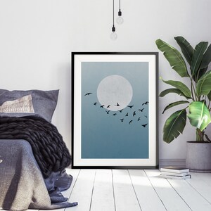Op de afbeelding: Een zwart ingelijst print met een blauwe en witte gradi&euml;nt achtergrond. Een grote witte cirkel bevindt zich in het midden van de print, met een zwerm zwarte vogels die er voor vliegen. De print hangt aan een witte muur boven een bed met een grijze deken.