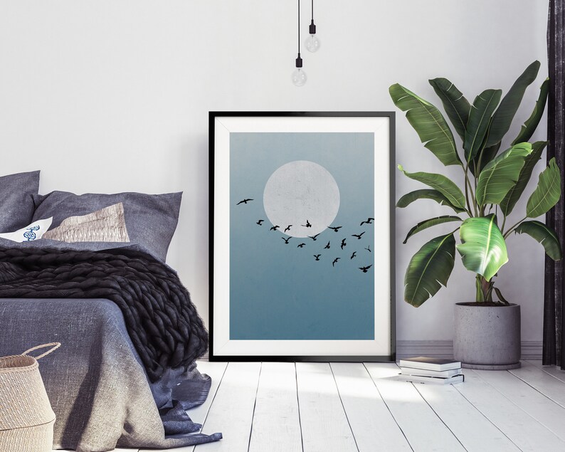 Op de afbeelding: Een zwart ingelijst print met een blauwe en witte gradi&euml;nt achtergrond. Een grote witte cirkel bevindt zich in het midden van de print, met een zwerm zwarte vogels die er voor vliegen. De print hangt aan een witte muur boven een bed met een grijze deken.