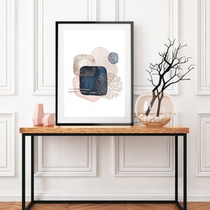 May include: A framed abstract art print with a white background and shapes in shades of pink, blue, and gold. The print is hanging on a white wall above a wooden console table with a vase of branches and two pink decorative objects.