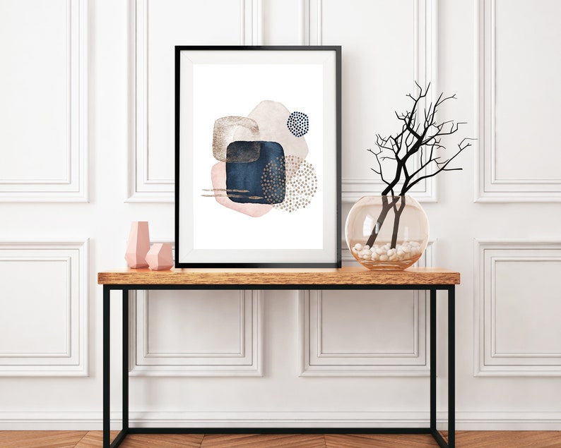 May include: A framed abstract art print with a white background and shapes in shades of pink, blue, and gold. The print is hanging on a white wall above a wooden console table with a vase of branches and two pink decorative objects.