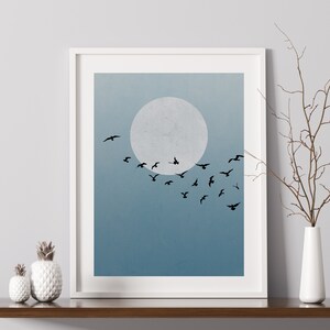 Op de afbeelding: Een ingelijst afdruk van een minimalistische illustratie van een zwerm vogels die vliegt voor een grote, witte maan tegen een blauwe lucht.