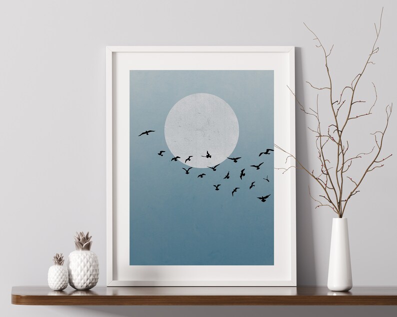 Op de afbeelding: Een ingelijst afdruk van een minimalistische illustratie van een zwerm vogels die vliegt voor een grote, witte maan tegen een blauwe lucht.