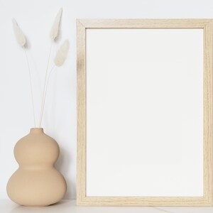 May include: A light brown ceramic vase with a rounded, bulbous shape, holding dried white pampas grass. A wooden picture frame with a white mat is on a white surface.
