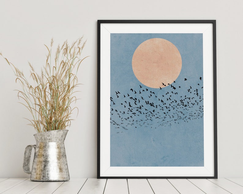 Pu&ograve; includere: Una stampa incorniciata di un design d'arte minimalista che presenta uno stormo di uccelli neri che volano in formazione contro un cielo blu con un grande cerchio arancione pallido al centro.