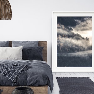 May include: A framed photograph of a misty forest scene with a white frame. The photograph is hanging on a white wall above a bed with a gray duvet cover and a woven throw blanket.