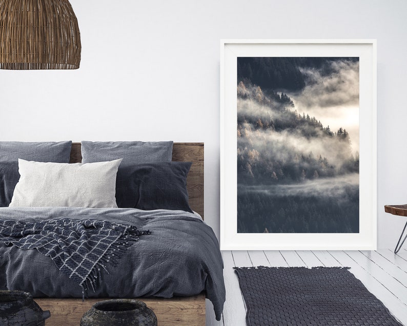 May include: A framed photograph of a misty forest scene with a white frame. The photograph is hanging on a white wall above a bed with a gray duvet cover and a woven throw blanket.