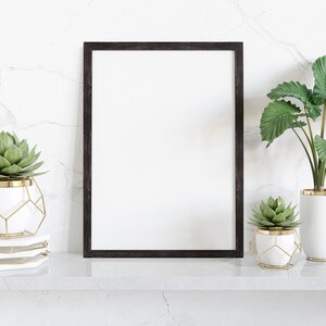 May include: A black framed poster mockup with a white background, sitting on a white marble shelf with two potted succulents. The succulents are in white pots with gold accents and geometric wire cages.