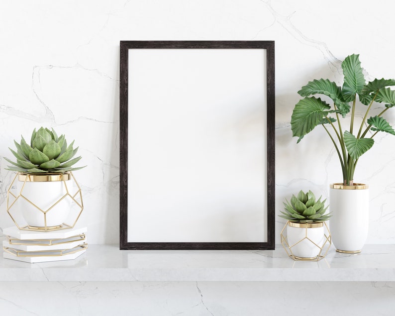 May include: A black framed poster mockup with a white background, sitting on a white marble shelf with two potted succulents. The succulents are in white pots with gold accents and geometric wire cages.