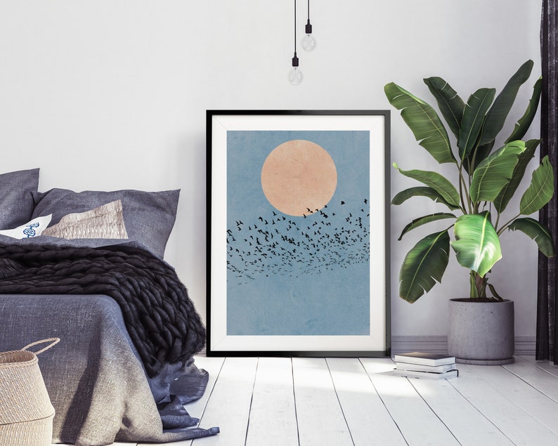 Pu&ograve; includere: Una stampa incorniciata di un'illustrazione minimalista di uno stormo di uccelli che vola davanti a un grande sole arancione pallido su uno sfondo di cielo blu.