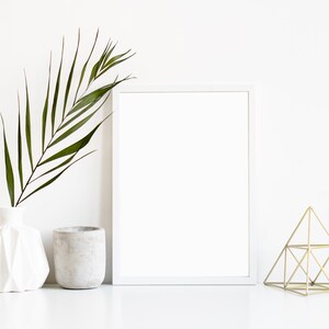 May include: A white frame with a blank canvas, a white ceramic vase with a green palm leaf, a gray concrete planter, and a gold geometric sculpture on a white surface.