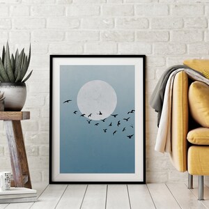 Op de afbeelding: Een zwart ingelijste print met een minimalistisch ontwerp in blauw en wit, met een volle maan en een zwerm vogels die in formatie vliegen.