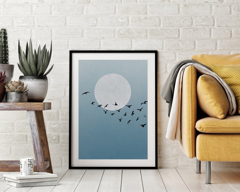 Op de afbeelding: Een zwart ingelijste print met een minimalistisch ontwerp in blauw en wit, met een volle maan en een zwerm vogels die in formatie vliegen.