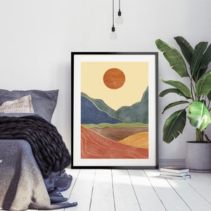 May include: A minimalist abstract landscape print with a large orange sun, blue and green mountains, and gold lines. The print is framed in black and hangs on a white wall.