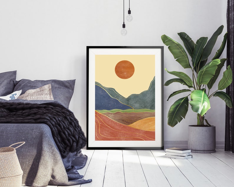 May include: A minimalist abstract landscape print with a large orange sun, blue and green mountains, and gold lines. The print is framed in black and hangs on a white wall.