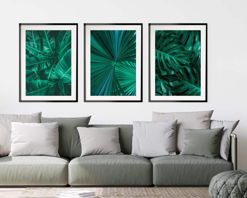 May include: Three framed prints of tropical leaves in shades of green. The prints are arranged in a row on a white wall above a gray sofa.