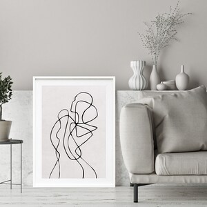 May include: A black and white line drawing of an abstract shape in a white frame on a white wall. The frame is on a white shelf with a small potted plant and a vase with dried flowers.