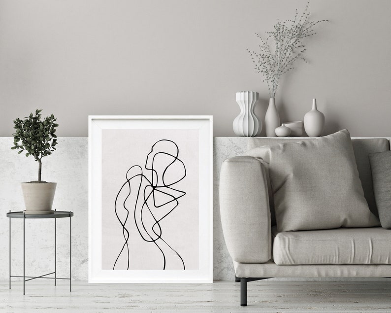 May include: A black and white line drawing of an abstract shape in a white frame on a white wall. The frame is on a white shelf with a small potted plant and a vase with dried flowers.