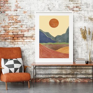 May include: A framed abstract landscape print with a yellow sun, blue and green mountains, and gold lines on a red, orange, and brown background. The print is hanging on a brick wall above a brown leather armchair with a black and white patterned pillow.