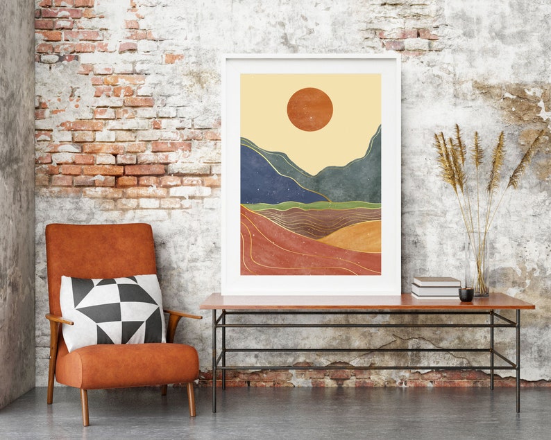 May include: A framed abstract landscape print with a yellow sun, blue and green mountains, and gold lines on a red, orange, and brown background. The print is hanging on a brick wall above a brown leather armchair with a black and white patterned pillow.