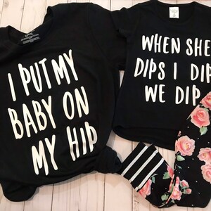 May include: Two black t-shirts with white text. The larger shirt says "I put my baby on my hip". The smaller shirt says "When she dips I dip we dip".