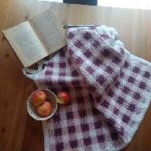 Puede incluir: Una manta tejida a cuadros blancos y burdeos, un libro abierto y un cuenco de manzanas sobre una mesa de madera. La escena evoca una sensación de comodidad y relajación, con un enfoque en el diseño de la manta.