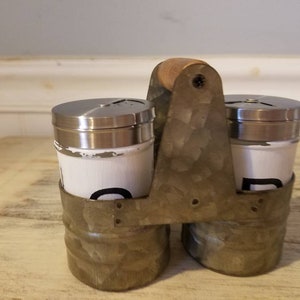 K&ouml;nnte beinhalten: Ein Set aus zwei wei&szlig;en Salz- und Pfefferstreuern mit schwarzer Beschriftung in einem verzinkten Metalltr&auml;ger mit einem Holzgriff.