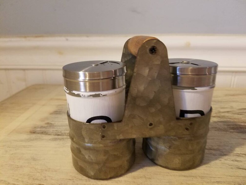K&ouml;nnte beinhalten: Ein Set aus zwei wei&szlig;en Salz- und Pfefferstreuern mit schwarzer Beschriftung in einem verzinkten Metalltr&auml;ger mit einem Holzgriff.