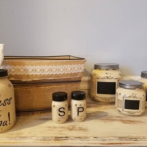 May include: A set of four cream-colored jars with black chalkboard labels. The jars are labeled "S", "P", and "Bless You!" and are arranged on a white wooden surface. Two smaller jars with black labels are also included, labeled "Mason".