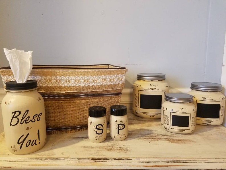 May include: A set of four cream-colored jars with black chalkboard labels. The jars are labeled "S", "P", and "Bless You!" and are arranged on a white wooden surface. Two smaller jars with black labels are also included, labeled "Mason".