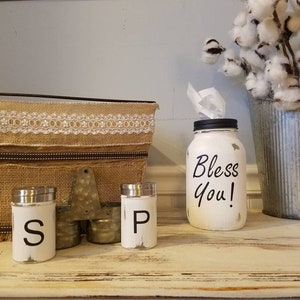 K&ouml;nnte beinhalten: Ein wei&szlig; bemaltes Einmachglas mit schwarzem Deckel und dem Text "Bless You!" auf der Vorderseite. Zwei wei&szlig; bemalte Salz- und Pfefferstreuer mit schwarzer Beschriftung. Ein Jutesack mit Spitzenbesatz und ein verzinkter Metalleimer mit Baumwollst&auml;ngeln.