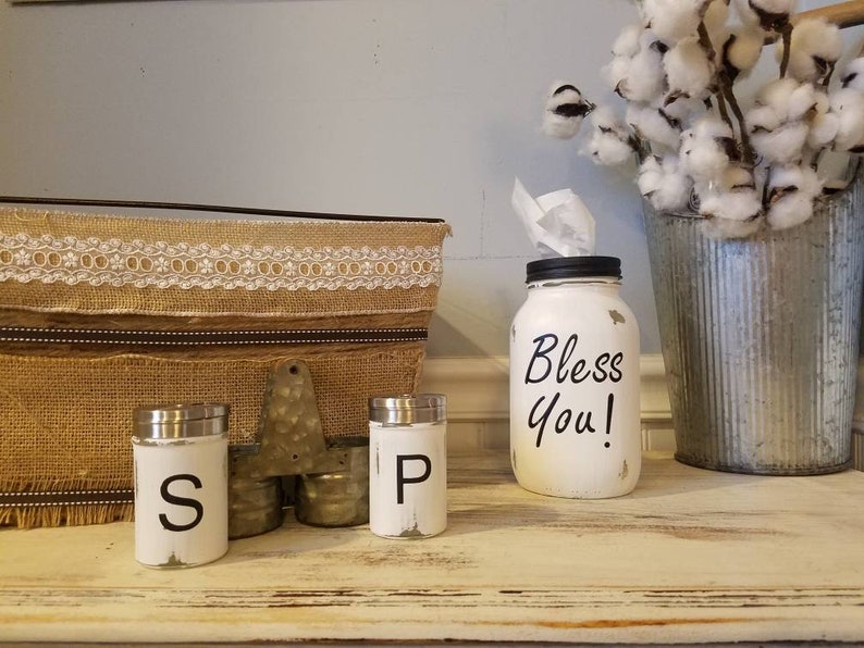 K&ouml;nnte beinhalten: Ein wei&szlig; bemaltes Einmachglas mit schwarzem Deckel und dem Text "Bless You!" auf der Vorderseite. Zwei wei&szlig; bemalte Salz- und Pfefferstreuer mit schwarzer Beschriftung. Ein Jutesack mit Spitzenbesatz und ein verzinkter Metalleimer mit Baumwollst&auml;ngeln.