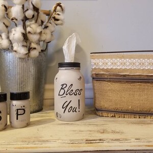 May include: A white painted mason jar with the words "Bless You!" written in black. The jar is filled with tissues and is sitting on a wooden table. There are two small mason jars with black lids and the letters "S" and "P" written on them. A burlap box with a white lace trim is also on the table.