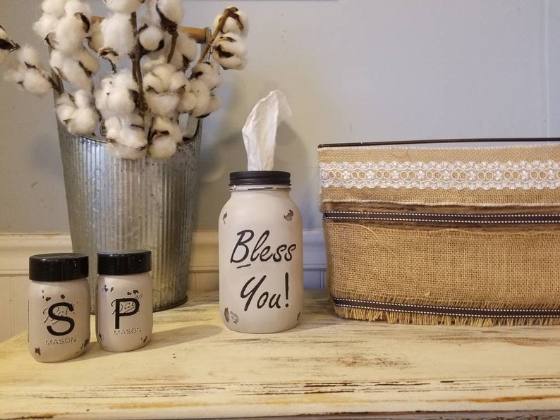 May include: A white painted mason jar with the words "Bless You!" written in black. The jar is filled with tissues and is sitting on a wooden table. There are two small mason jars with black lids and the letters "S" and "P" written on them. A burlap box with a white lace trim is also on the table.