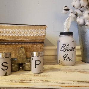 K&ouml;nnte beinhalten: Ein wei&szlig; bemaltes Einmachglas mit schwarzem Deckel und den Worten "Bless You!" in Schwarz. Das Glas wird als Taschentuchspender verwendet. Es gibt zwei wei&szlig; bemalte Salz- und Pfefferstreuer aus Metall mit schwarzen Buchstaben. Die Streuer stehen auf einer Holzoberfl&auml;che mit einer Kiste aus Sackleinen und Spitze dahinter.