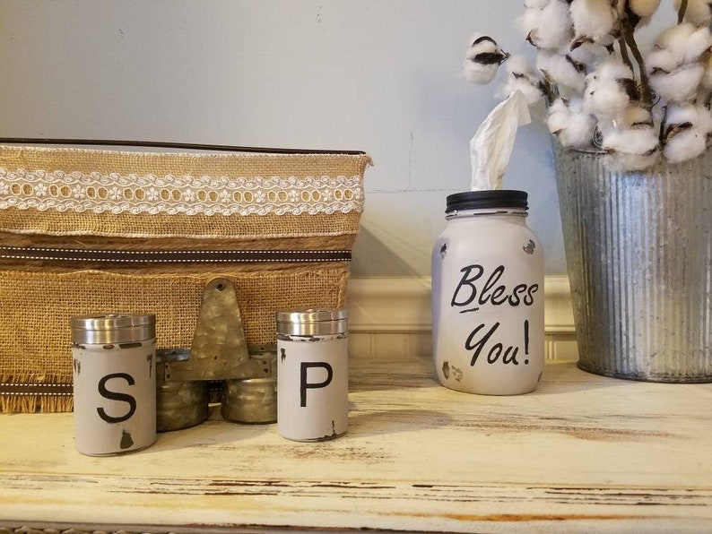 K&ouml;nnte beinhalten: Ein wei&szlig; bemaltes Einmachglas mit schwarzem Deckel und den Worten "Bless You!" in Schwarz. Das Glas wird als Taschentuchspender verwendet. Es gibt zwei wei&szlig; bemalte Salz- und Pfefferstreuer aus Metall mit schwarzen Buchstaben. Die Streuer stehen auf einer Holzoberfl&auml;che mit einer Kiste aus Sackleinen und Spitze dahinter.