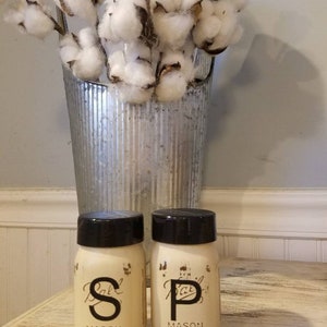 May include: Two white painted mason jars with black lids and black lettering. The jars are labeled "Salt" and "Pepper" and are sitting on a wooden table. A vase with cotton stems is in the background.