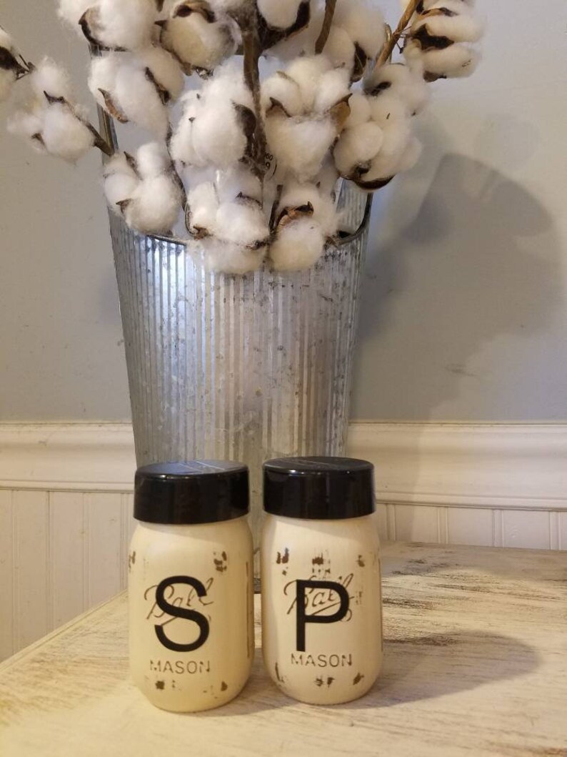May include: Two white painted mason jars with black lids and black lettering. The jars are labeled "Salt" and "Pepper" and are sitting on a wooden table. A vase with cotton stems is in the background.