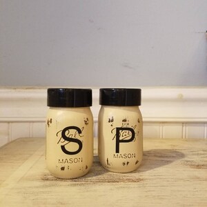 May include: Two vintage-style glass salt and pepper shakers with black lids. The shakers are labeled "S" and "P" with the word "MASON" underneath. The shakers are sitting on a light wood surface.